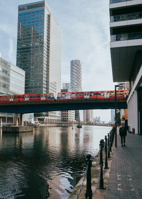 The image shows a river running through an urban area with modern high-rise buildings on either side. A red and white train is crossing a bridge over the river, situated above the water and supported