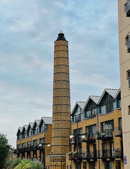 A tall, narrow brick chimney with black metal bands running horizontally around it, standing in front of modern residential buildings with multiple balconies decorated with potted plants and outdoor f