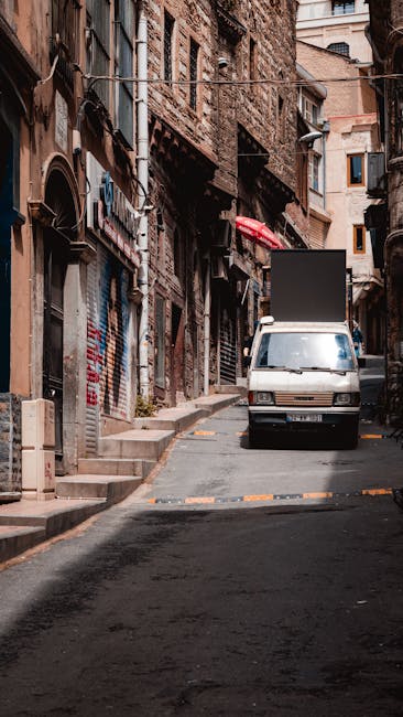 A narrow residential street in Limehouse with a white moving van parked on the pavement, partially blocking the sidewalk. The van's rear doors are open, indicating active loading or unloading. The surrounding buildings are old, multi-storey brick structures with small windows, some featuring external air conditioning units and street-level shops with metal shutters and signage. Various cardboard boxes wrapped in plastic, along with furniture pieces covered in blankets and plastic wrap, are visible near the van, suggesting a home relocation process. Metal trolleys and straps are on the ground, being used to facilitate moving heavy items through the constrained street space typical of house removals in densely built environments. Natural daylight illuminates the scene, highlighting the urban setting and the logistical challenges of furniture transport in narrow streets, illustrating the professional removals services provided by Man and Van Docklands.