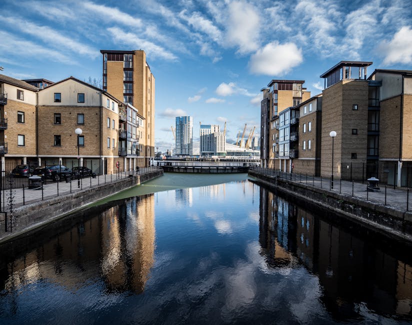 View of a narrow waterway in Limehouse with modern residential buildings on either side, featuring brick facades, small balconies, and large windows. The water reflects the buildings and the partly cloudy blue sky above. In the background, contemporary high-rise office towers with glass exteriors are visible, along with construction cranes indicating ongoing development. A bridge crosses over the waterway, and the pavement along the edge has black metal railings, with street lamps and parked cars nearby. The scene captures an urban riverside setting typical for house removals and furniture transport within the Docklands area, highlighting the close proximity between residential properties and transport routes for home relocation projects managed by Man and Van Docklands, especially on narrow streets suitable for specialist moving services and packing and moving logistics.