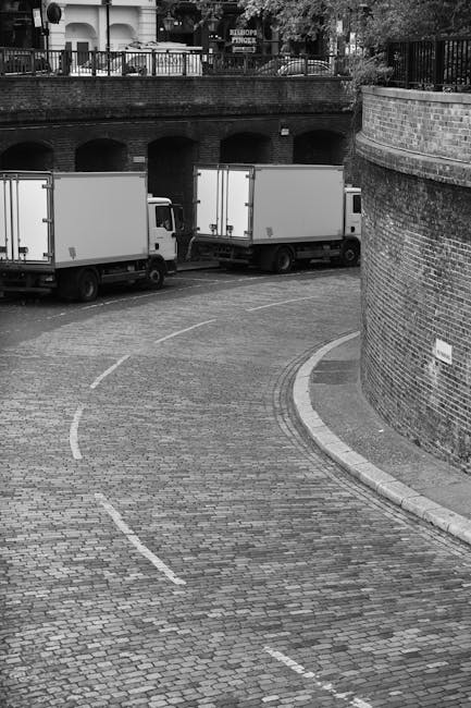 A narrow residential street in Limehouse with a white moving van parked on the pavement, partially blocking the sidewalk. The van's rear doors are open, indicating active loading or unloading. The surrounding buildings are old, multi-storey brick structures with small windows, some featuring external air conditioning units and street-level shops with metal shutters and signage. Various cardboard boxes wrapped in plastic, along with furniture pieces covered in blankets and plastic wrap, are visible near the van, suggesting a home relocation process. Metal trolleys and straps are on the ground, being used to facilitate moving heavy items through the constrained street space typical of house removals in densely built environments. Natural daylight illuminates the scene, highlighting the urban setting and the logistical challenges of furniture transport in narrow streets, illustrating the professional removals services provided by Man and Van Docklands.