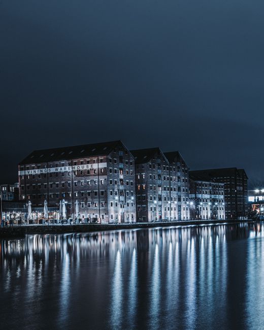 A nighttime scene showing a row of residential or commercial buildings with multiple illuminated windows next to a waterfront. The ground floor has an arcade-style structure with open arches, behind w